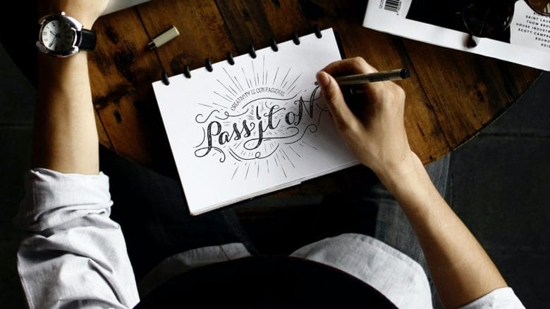 Person writing Passion in decorative lettering on a notepad at a wooden table with a watch, book, and other items visible.