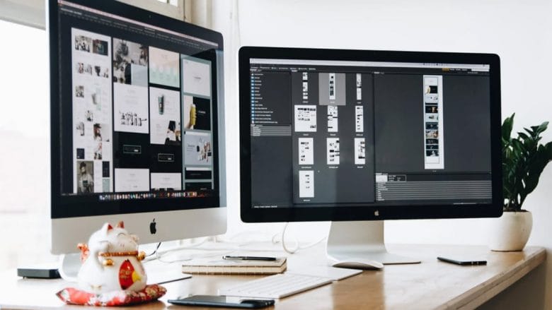 Two computer monitors on a desk displaying graphic design software, accompanied by a notebook, a smartphone, a pen, and a small figurine.