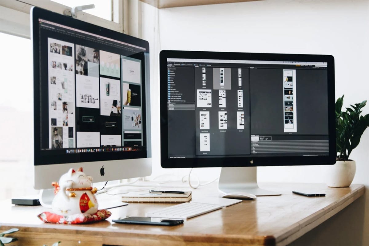 Two computer monitors on a desk displaying graphic design software, accompanied by a notebook, a smartphone, a pen, and a small figurine.