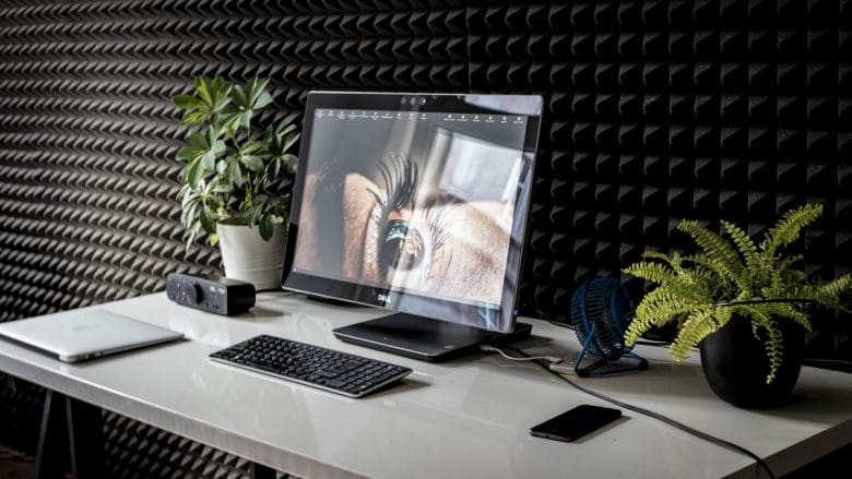 A modern workspace featuring a laptop with a secondary monitor, a wireless keyboard, a notebook, a smartphone, and two potted plants on a white desk against a textured black wall.