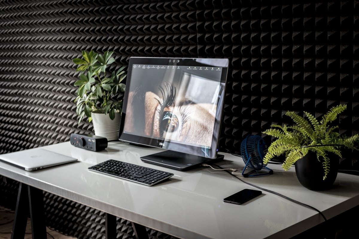 A modern workspace featuring a laptop with a secondary monitor, a wireless keyboard, a notebook, a smartphone, and two potted plants on a white desk against a textured black wall.
