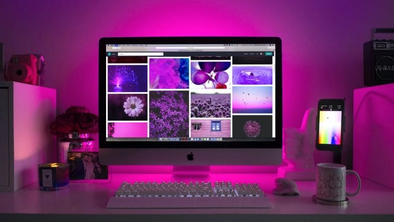 A well-organized desk with an imac displaying a collage of purple-themed images, flanked by a smartphone in a holder, a coffee mug, and various desk accessories, all under ambient purple lighting.