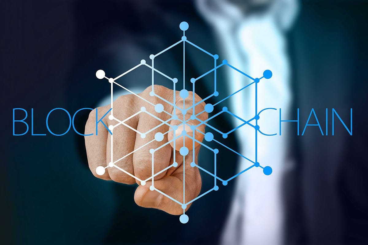 A person in a suit points at a blockchain graphic with interconnected nodes, with the word BLOCKCHAIN displayed in the background.