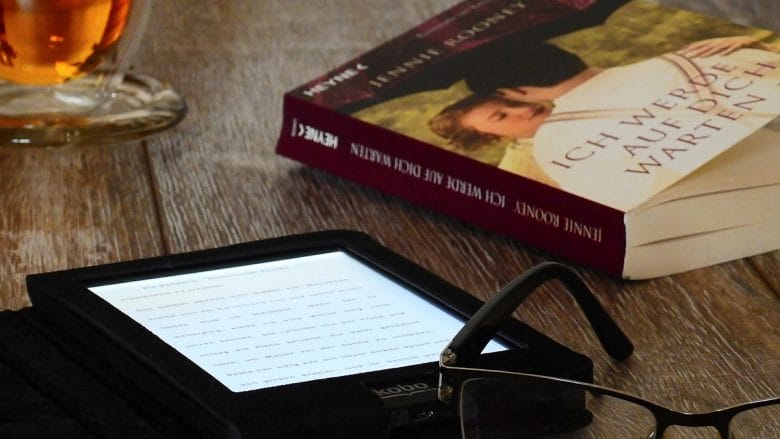 A hardcover book, e-reader, eyeglasses, and a cup of tea arranged on a wooden surface.