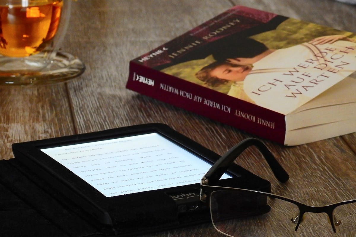 A hardcover book, e-reader, eyeglasses, and a cup of tea arranged on a wooden surface.