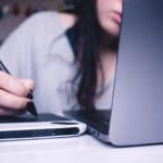A woman writing on a laptop with a pen.