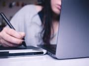 A woman writing on a laptop with a pen.