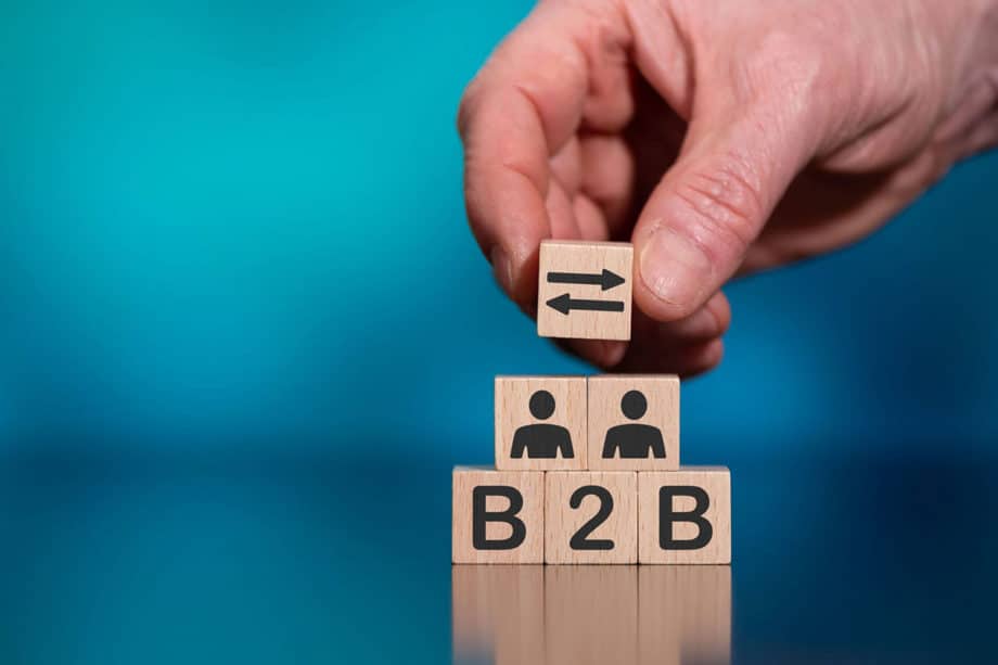 A hand placing a wooden block with a left arrow symbol on top of three blocks stacked vertically that display "b2b" and two icons representing people, against a blue background.