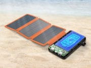 A solar charger with four foldable panels and a digital display is placed on sandy beach near the water.