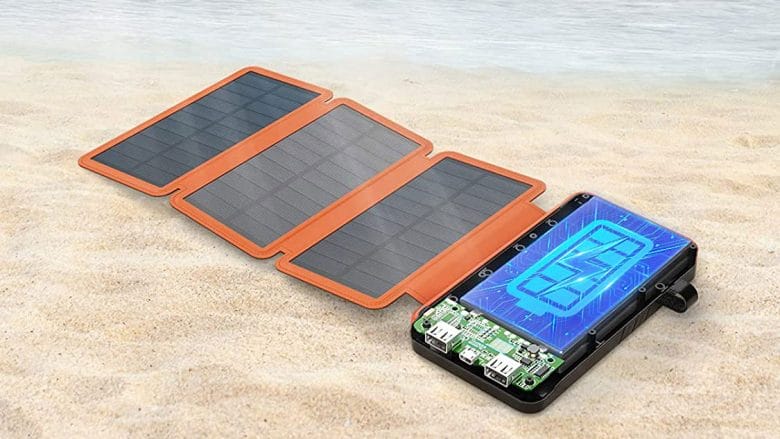 A solar charger with four foldable panels and a digital display is placed on sandy beach near the water.