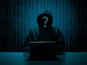 A hooded figure at a laptop with a question mark over their face, set against a backdrop of digital numbers.