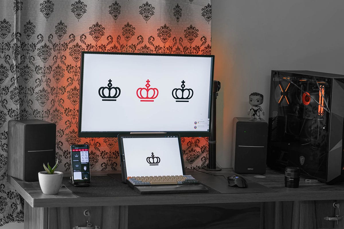 A modern computer setup on a wooden desk featuring a large monitor with a triple crown graphic wallpaper, a mechanical keyboard, and a smartphone on a stand, with a gaming pc tower and speakers on the sides.