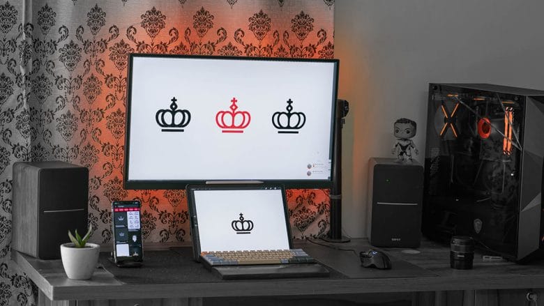 A modern computer setup on a wooden desk featuring a large monitor with a triple crown graphic wallpaper, a mechanical keyboard, and a smartphone on a stand, with a gaming pc tower and speakers on the sides.