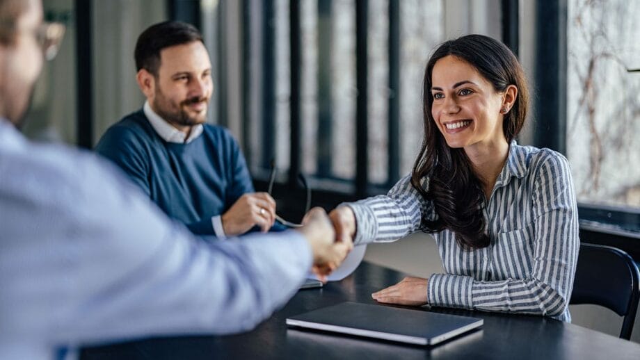 Two professionals are shaking hands across a table, likely concluding a meeting or agreement, while a third individual observes the interaction with a smile.