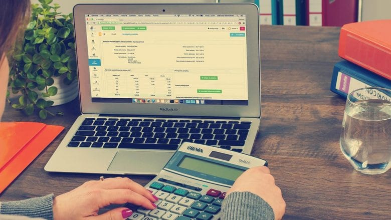 Person using a calculator in front of an open laptop displaying a spreadsheet. A glass of water and office supplies are on the wooden desk.