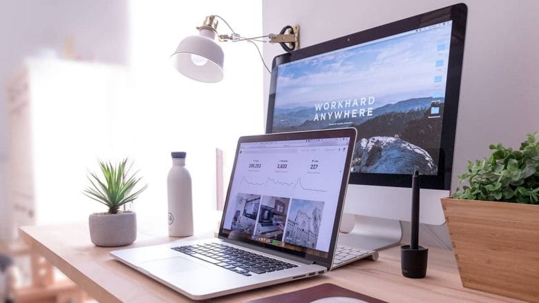 A modern workspace featuring an imac and macbook on a wooden desk, accompanied by a potted plant and a white water bottle, illuminated by a desk lamp.