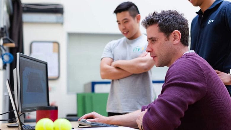 Three men in a sports-themed setting observe a computer screen. One is seated and holding a mouse, while the others stand nearby with interest.