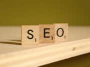 Three wooden Scrabble tiles spell out "SEO" on a wooden surface against a green background.