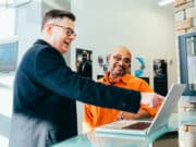 Two men stand at a glass counter, one pointing at a laptop screen while the other listens and smiles.