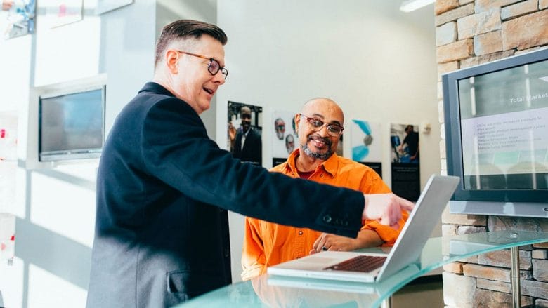 Two men stand at a glass counter, one pointing at a laptop screen while the other listens and smiles.