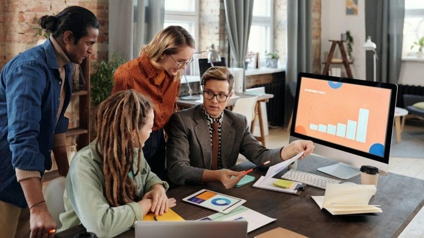 Four people discuss documents at a desk with charts and a computer displaying a bar graph in a modern office setting.