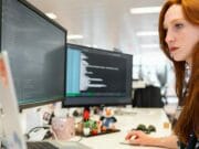 A woman with red hair works at a desk with two large computer monitors displaying lines of code in an office setting.