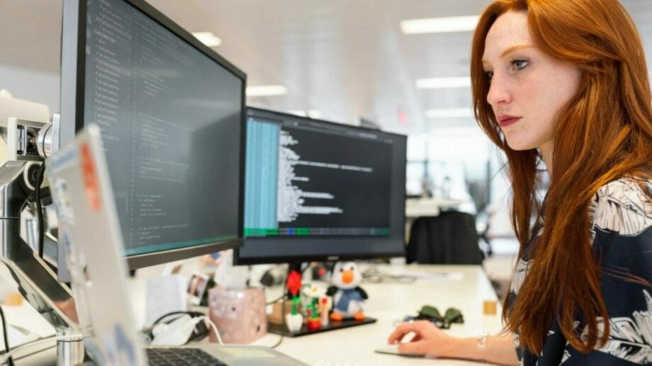 A woman with red hair works at a desk with two large computer monitors displaying lines of code in an office setting.