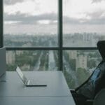 A man sitting at a desk with a laptop in front of a window.