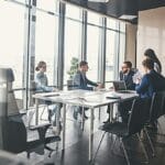 Five people in business attire sit and stand around a conference table in a modern office meeting room with large windows.