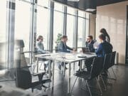 Five people in business attire sit and stand around a conference table in a modern office meeting room with large windows.