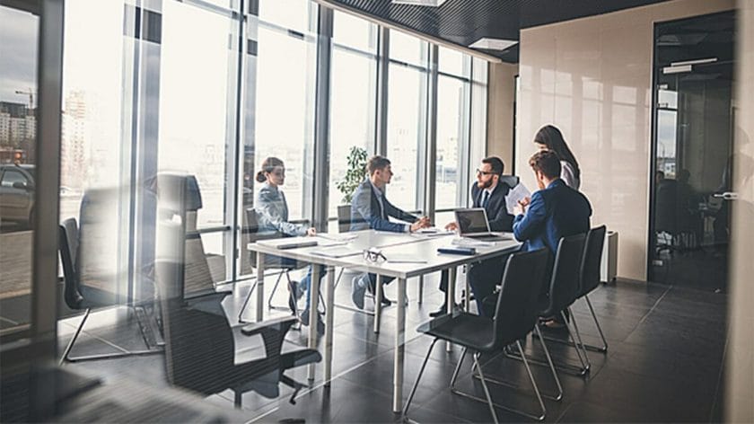 Five people in business attire sit and stand around a conference table in a modern office meeting room with large windows.