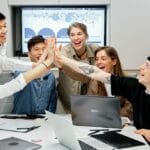 Six people sitting at a conference table high-five each other, smiling, with laptops and papers in front of them.