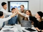 Six people sitting at a conference table high-five each other, smiling, with laptops and papers in front of them.