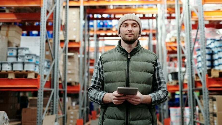 A man wearing a beanie and puffer vest holds a tablet while standing in a warehouse aisle with shelves stocked with boxes and containers.