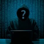 A hooded figure at a laptop with a question mark over their face, set against a backdrop of digital numbers.