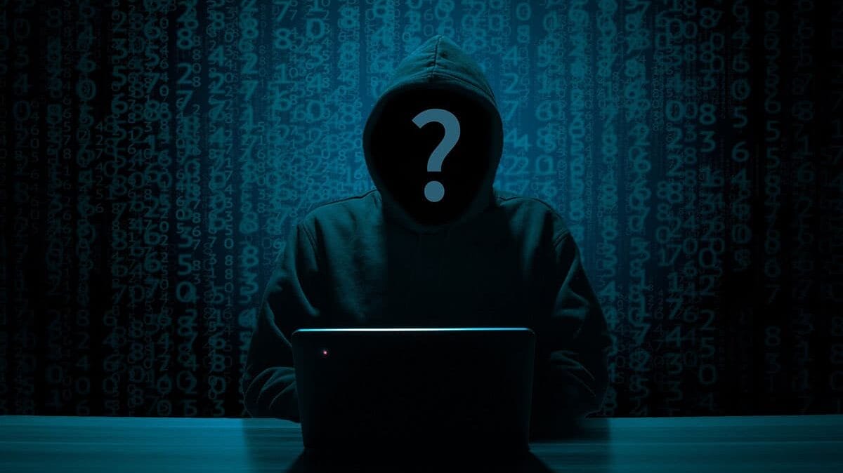A hooded figure at a laptop with a question mark over their face, set against a backdrop of digital numbers.