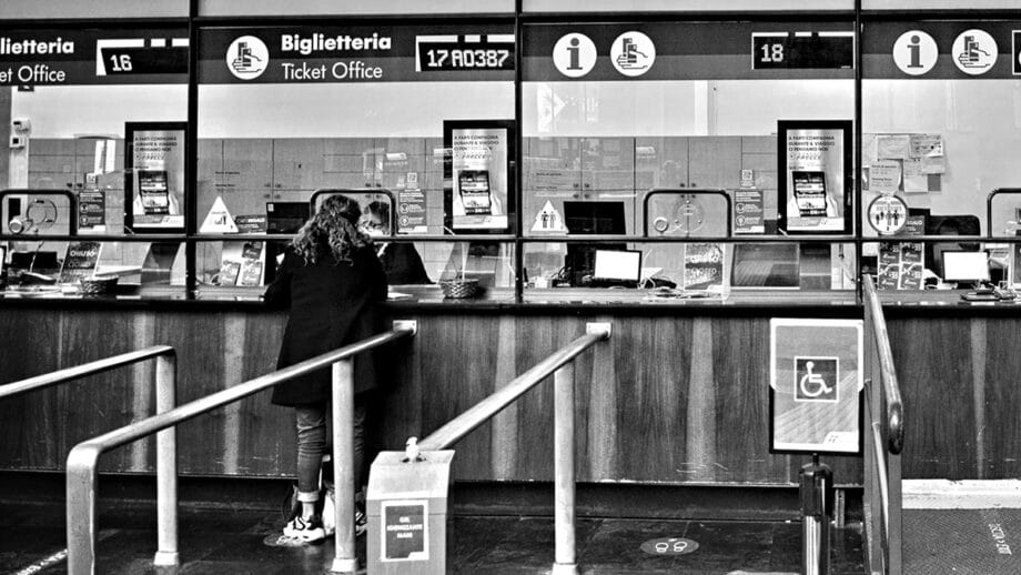 A person standing at a ticket office with multiple service windows, information signs, and an accessible entrance sign visible.
