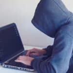A person in a gray hoodie working on a laptop showing lines of code, seated at a white desk.