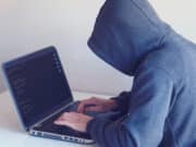 A person in a gray hoodie working on a laptop showing lines of code, seated at a white desk.