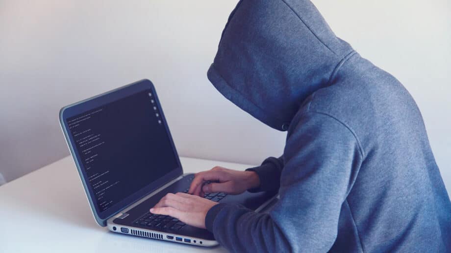 A person in a gray hoodie working on a laptop showing lines of code, seated at a white desk.