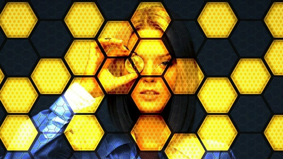 A woman holds a coin up to her eye, seen through a honeycomb-patterned overlay with yellow and black hexagons.