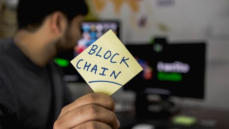 A person holds a yellow sticky note with BLOCK CHAIN written on it, with computer monitors visible in the background.