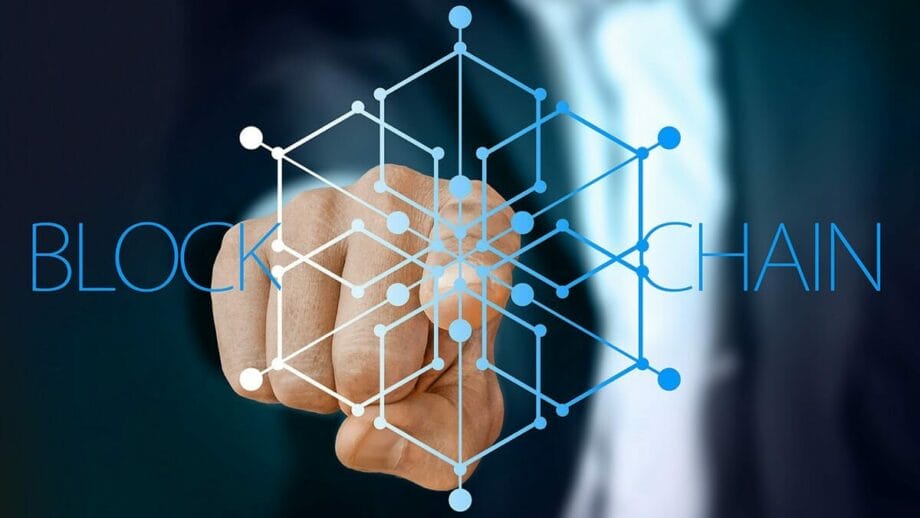 A person in a suit points at a blockchain graphic with interconnected nodes, with the word BLOCKCHAIN displayed in the background.