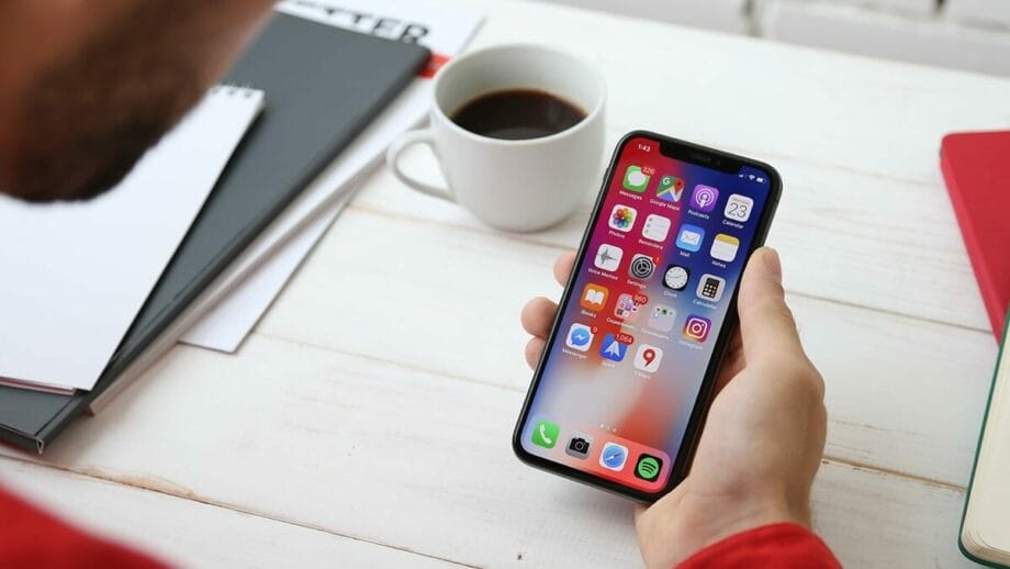 Person holding a smartphone displaying multiple app icons, with a coffee cup and notebooks on a table.