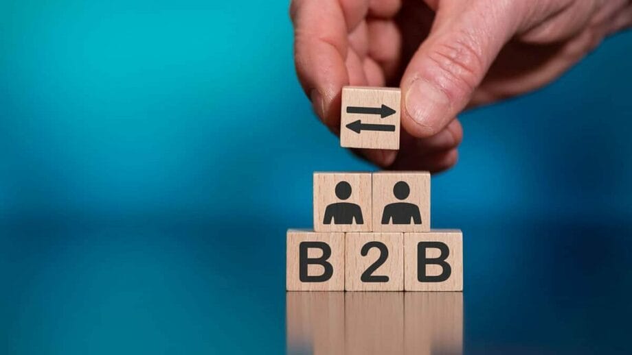 A hand placing a wooden block with a left arrow symbol on top of three blocks stacked vertically that display "b2b" and two icons representing people, against a blue background.