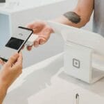 A person pays at a checkout counter using a smartphone and Google Pay, while another person holds a point-of-sale terminal.