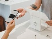 A person pays at a checkout counter using a smartphone and Google Pay, while another person holds a point-of-sale terminal.