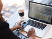 Person working at a round table with a laptop showing code, a smartphone connected by cable, and a reusable coffee cup nearby.