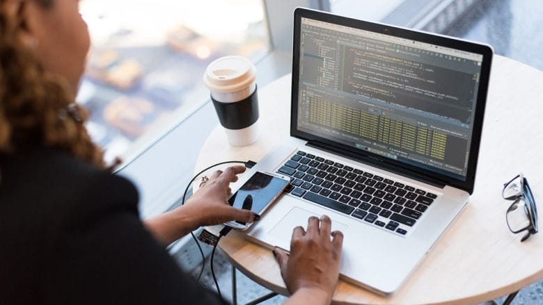 Person working at a round table with a laptop showing code, a smartphone connected by cable, and a reusable coffee cup nearby.