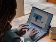 Person types on a laptop at a wooden table with a mug nearby, browsing a website displaying a man in a winter jacket.
